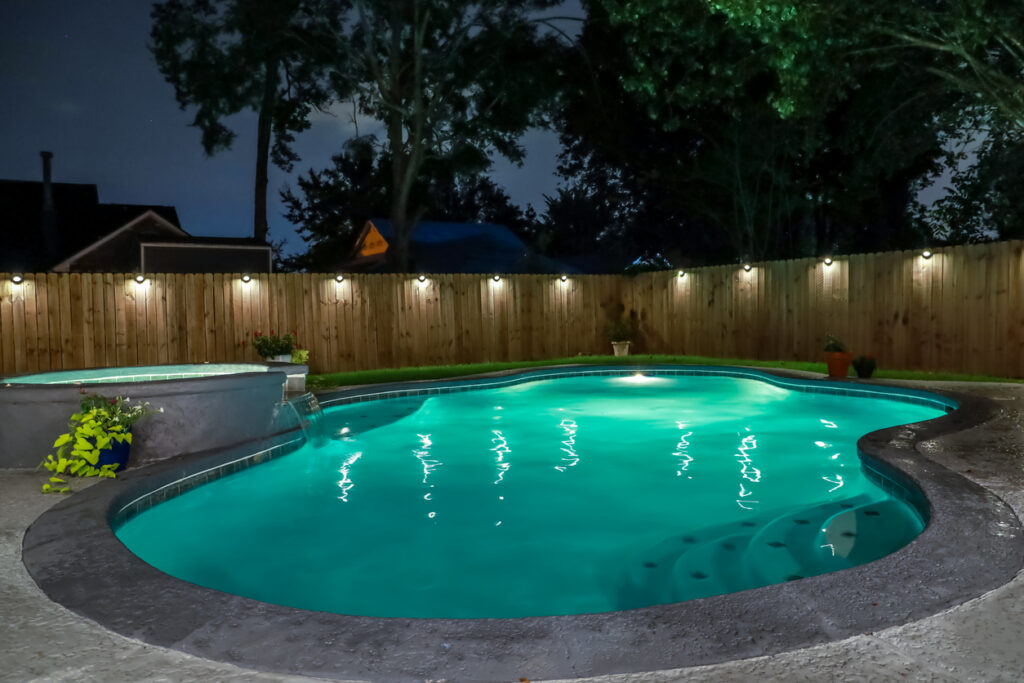 A backyard swimming pool and hot tub at night with solar lights around the fence for privacy and illumination.
