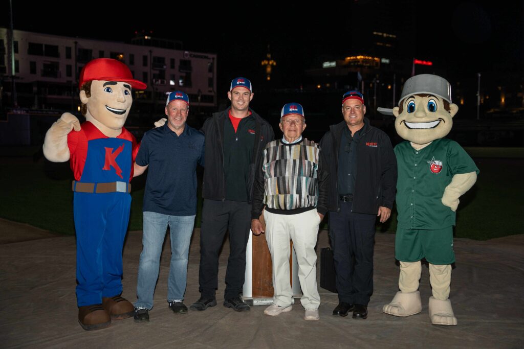 Group of five men at a baseball field during nighttime. Two are in mascot costumes, while the others wear casual outfits and caps, smiling warmly.