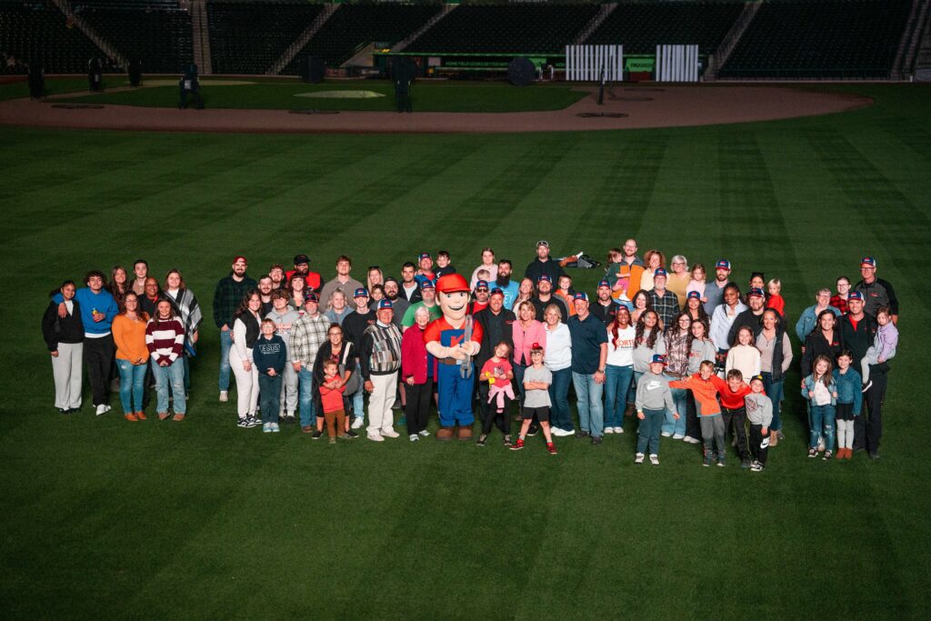 A large group of people poses for a photo on a baseball field with a person in a team mascot costume standing in the center.