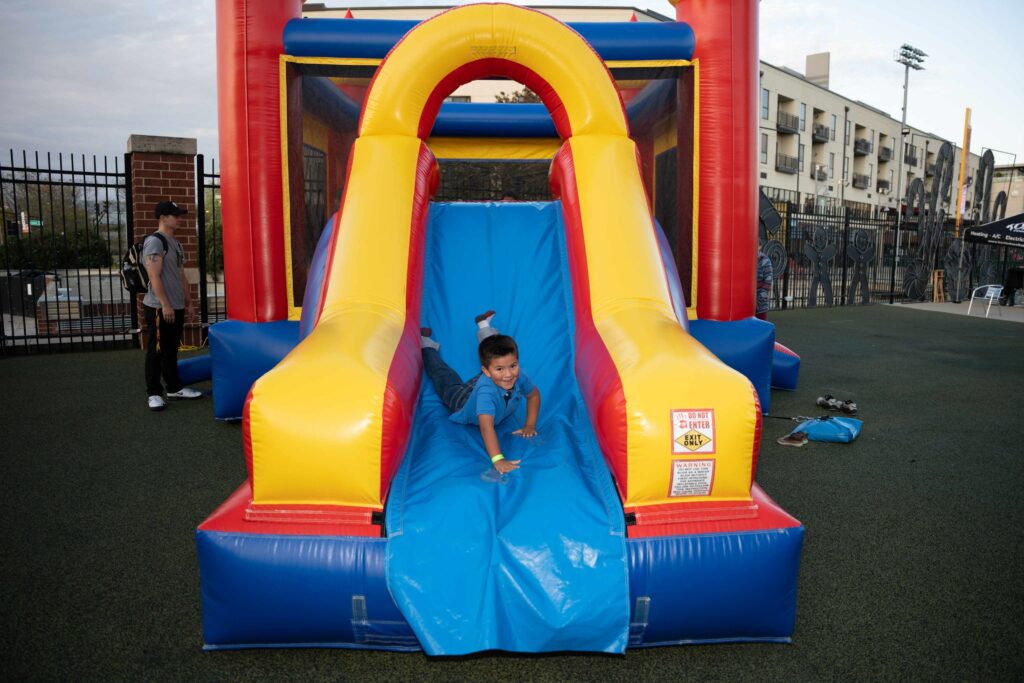 A child joyfully slides down a bright yellow and blue inflatable slide. The scene is lively and playful, set outdoors with a fenced background.