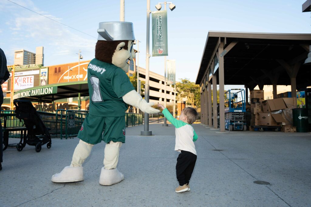 Child joyfully holds hands with a mascot dressed in green sports attire and helmet, set in an outdoor stadium entrance. Warm lighting creates a friendly atmosphere.